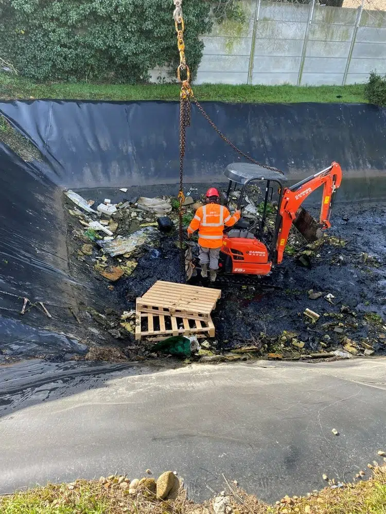 Photo d'un nettoyage en milieu industriel. Bassin de rétention d'eau avec un salarié et une mini pelle.